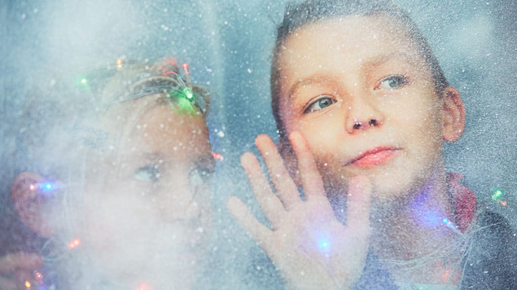 Close up of young brother and sister wrapped in Christmas lights and looking out a frosted window