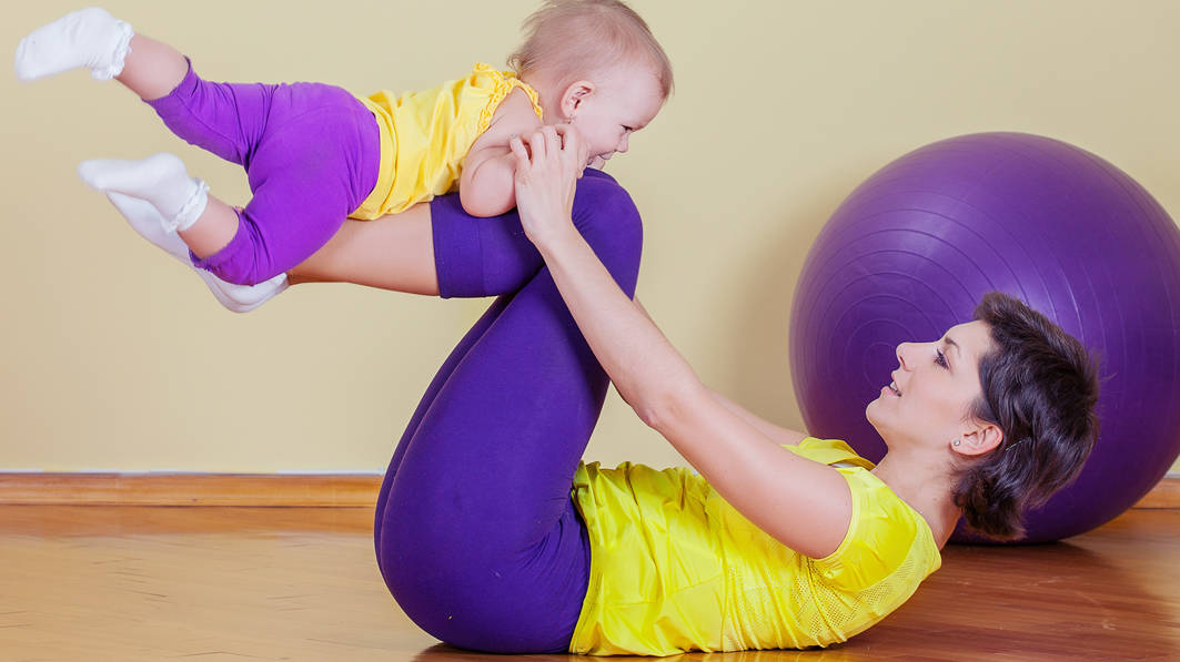 Young mom in yellow shirt and purple tights lying on floor, balancing toddler on her legs who’s dressed in a similar outfit