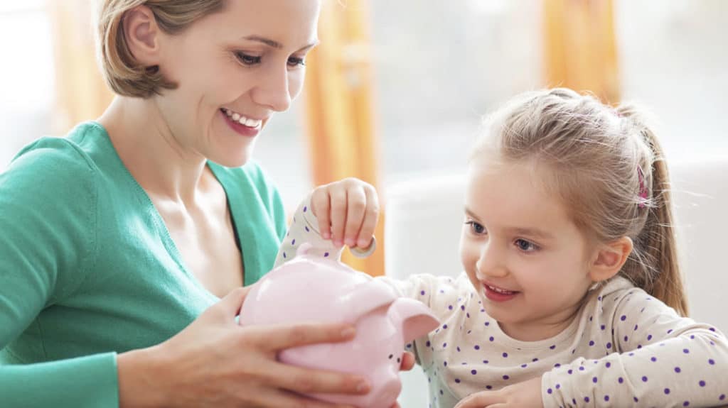 In teaching kids about money, smiling mom holding a piggy bank with her young daughter adding a coin to it