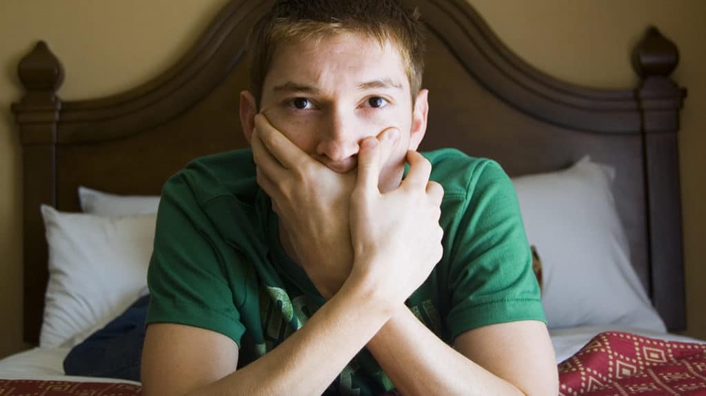 Teen boy sitting on the edge of his bed with his hands cupped over his mouth, looking like he’s troubled or shocked