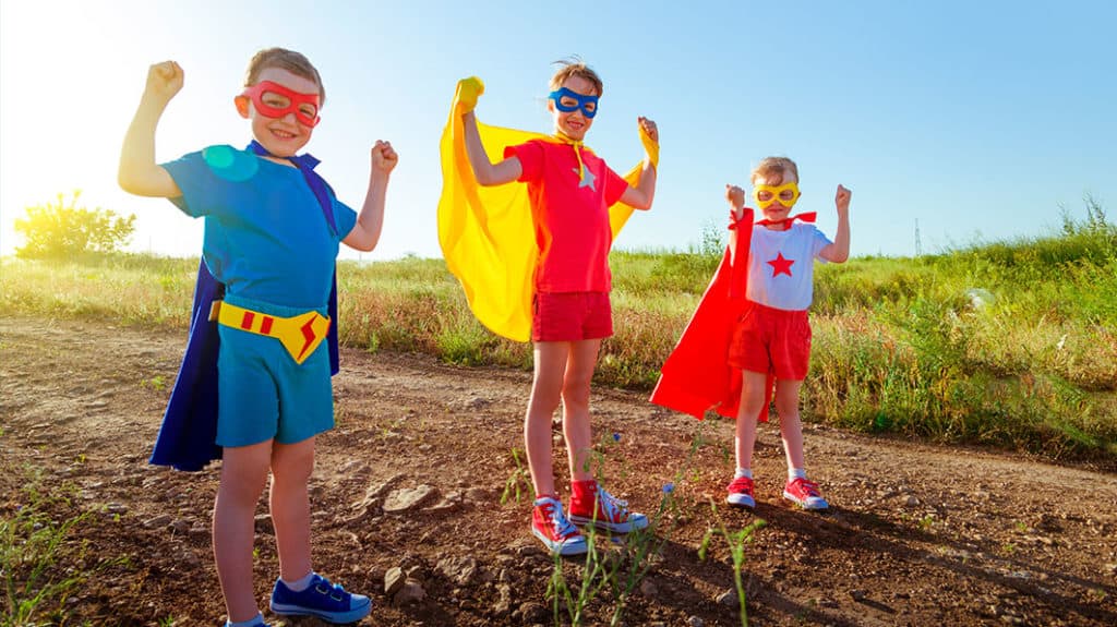 Three smiling kids standing on a dirt road on a sunny day wearing super hero costumes with capes and flexing their biceps