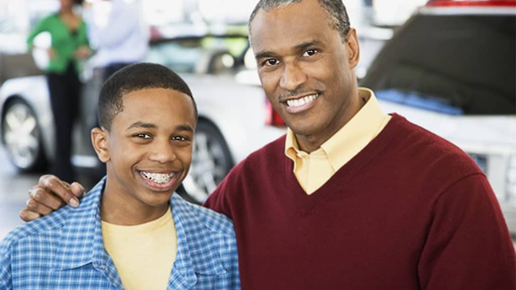 Smiling father and teen son posing for the camera, with a white car and two people in a blurred out background.