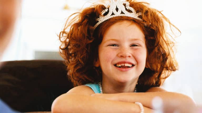 Young smiling red-headed girl with a princess crown and her arms folded/resting on a table
