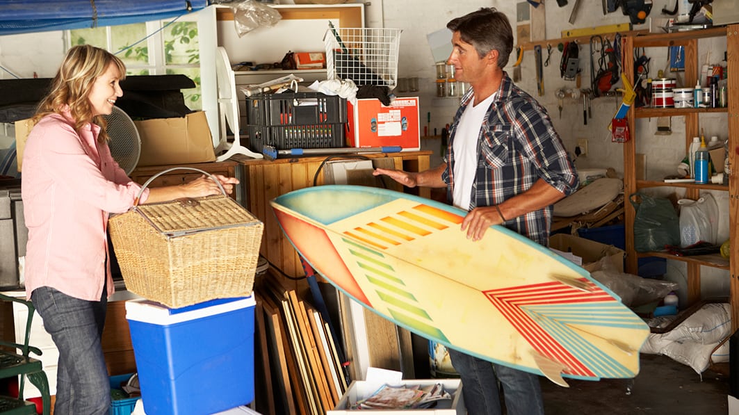 Couple standing and talking in their junk-filled garage. He's holding a surfboard, her arms are resting on a picnic basket.