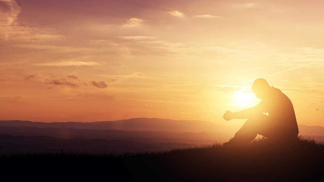 Silhouetted profile of man sitting and praying on a hillside with the setting sun shining directly behind him.
