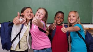 Group of four smiling kids with backpacks standing in front of a chalkboard. All are giving a thumbs-up to the camera.