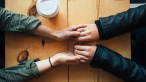 Shown from above, a married couple's hands holding each other while resting on a table with a cup of coffee to the side.