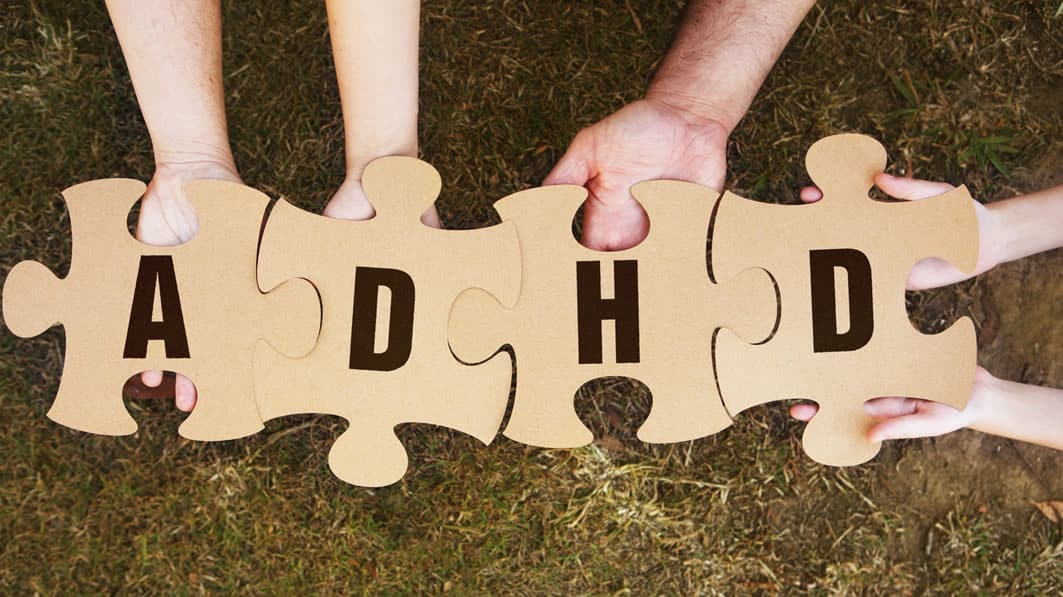 Shown from above, hands holding large puzzle pieces, each piece having a letter on it, together spelling out ADHD.