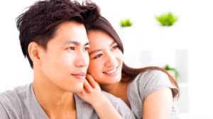 Profile of happy, young Asian couple looking at something off camera. She's resting her hands and head on his shoulders.