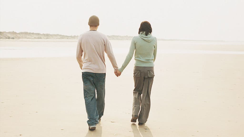 Shown from behind, a couple dressed for cool weather holding hands and walking down the beach.