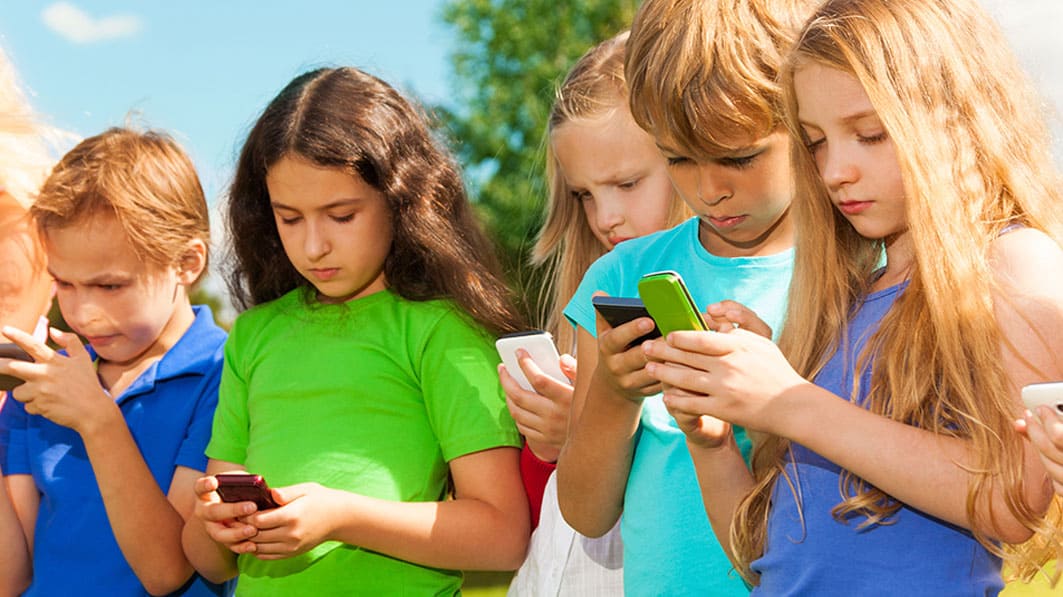 Colorful photo of small group of young children looking intently at their cell phones while standing outside on a sunny day.