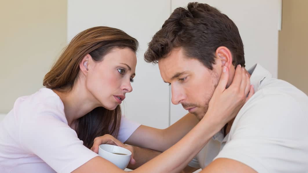 Seated young couple. He's looking downward and depressed. She's looking at him worriedly, her hands wrapped around his face.