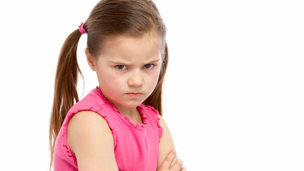 Close up of young girl against a white background. She's looking into the camera angrily, her arms folded across her chest.