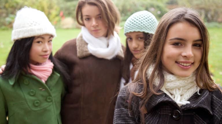 Four teen girls posing outside on a Fall day. One stands separately in the foreground smiling, the others look disapproving.