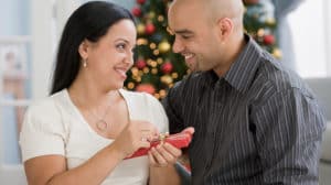 Smiling couple with Christmas tree in the background. She's about to open a small gift from him.