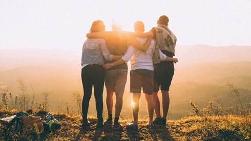 Shown from behind, a group of four people, their arms around each other as they watch a sunrise over the mountains