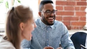 Young man and woman sitting at a table in a meeting. They’re both smiling and laughing.