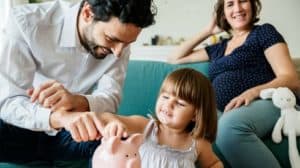 Father and young daughter putting money in a piggybank, with pregnant mom in the background