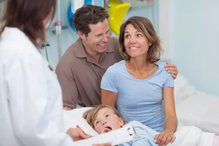 Happy parents receiving good news from a doctor about their daughter who’s lying next to them in a hospital bed after a health scare.