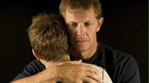 Somber father hugging teen son with black background