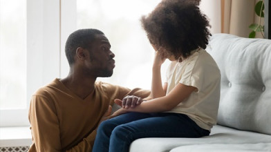 Concerned dad kneeling beside and trying to comfort his sad young daughter who's sitting on a couch