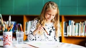 Young girl seated at a table poised to paint with a brush in her hand. A jar of additional brushes sits beside her.