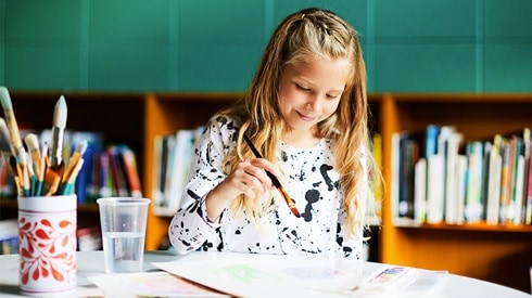 Young girl seated at a table poised to paint with a brush in her hand. A jar of additional brushes sits beside her.