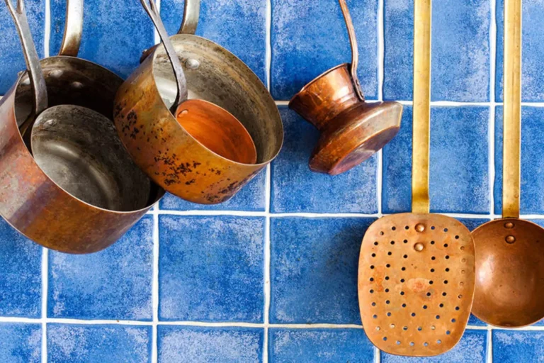 A collection of copper cookware with visible scuff marks hanging against a vibrant blue tiled wall, illustrating the beauty of wear and utility.