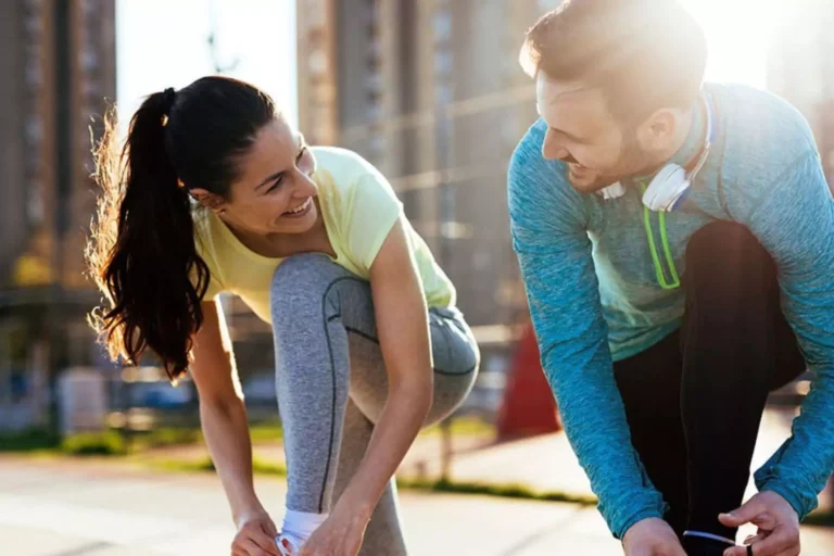 A couple tying their shoelaces and smiling at each other, symbolizing the role of self-care in maintaining a healthy and supportive relationship.