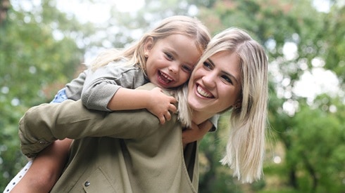 Mom giving her young daughter a piggyback ride outside; both are smiling happily