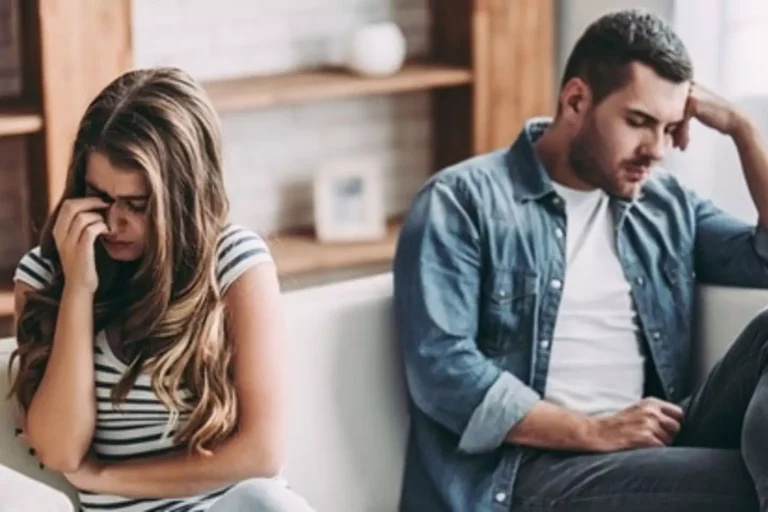 Young couple sitting on a couch experiencing relationship problems. He's downcast, she's turned away from him crying.