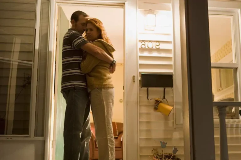 A somber-looking husband and wife hugging on their porch in the evening, consoling each other during a difficult time