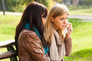 Two women sitting on a park bench, one visibly upset and holding a tissue, while the other offers quiet support, symbolizing challenges like how to stay married when your friends are divorcing.
