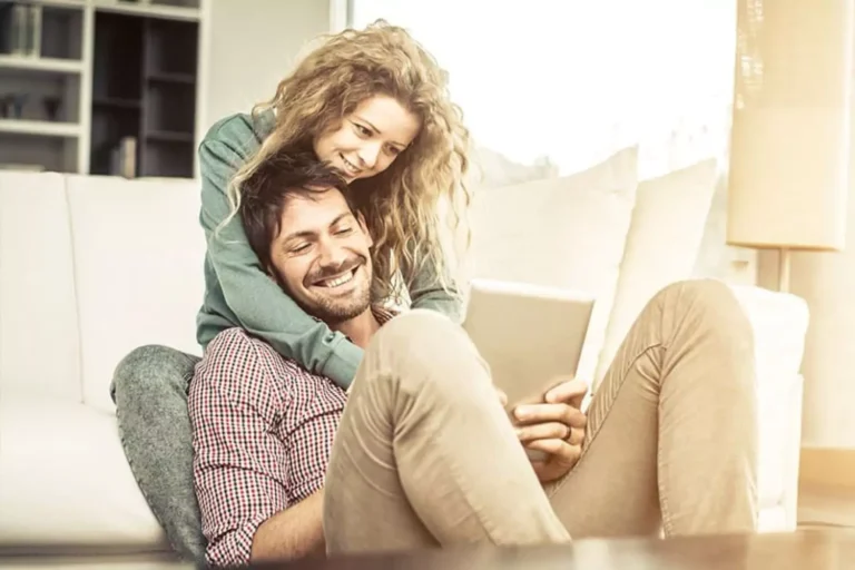 A smiling couple relaxing at home, the wife leaning affectionately on her husband’s shoulder while he holds a tablet, symbolizing the importance of the phrase “if you want a better marriage, listen to your wife.”
