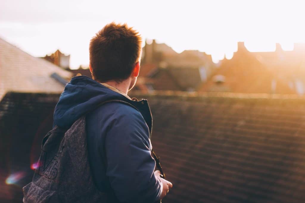 Young man with a backpack standing on a roof looking away from the camera over neighborhood rooftops