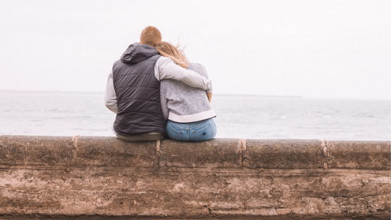 Shown from behind, a man and woman sitting on a low wall overlooking the ocean. He has his arm around her.
