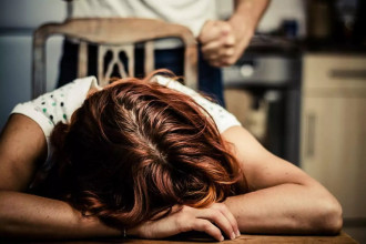 A distressed woman lays her head on a table while an abusive spouse stands behind her with a clenched fist.