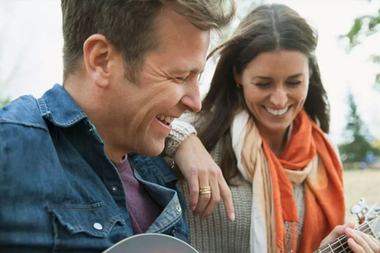 A couple, symbolizing Ron and Opal Erickson, laughing together outdoors while playing a guitar, enjoying a lighthearted moment.