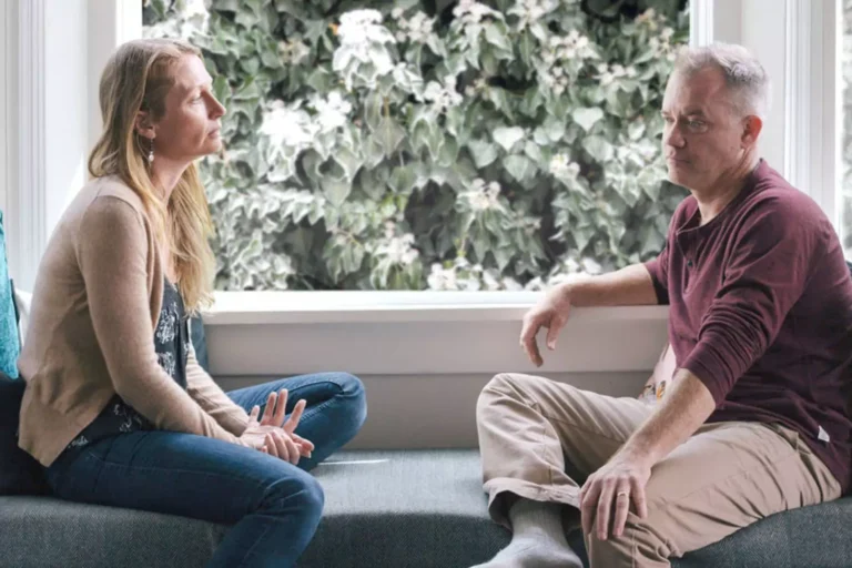A middle-aged couple sits on a couch facing each other, engaged in a serious conversation about staying married, with expressions of concern and contemplation.