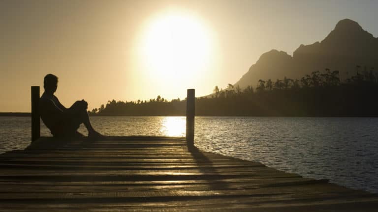 Silhouette of a man sitting on a dock looking out over a lake at sunset