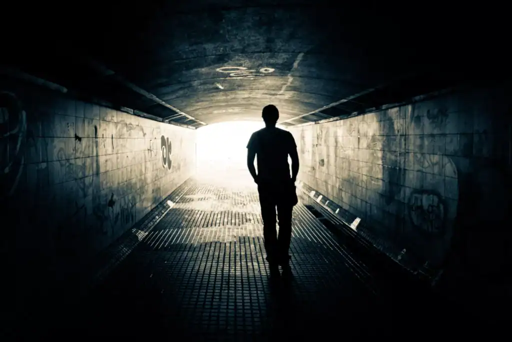 A silhouette of a person walking through a dimly lit tunnel toward a bright light at the end.