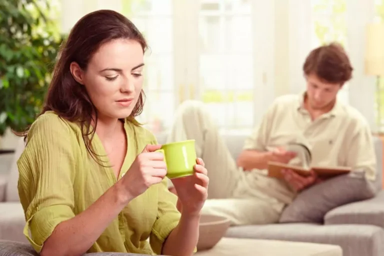 Woman in green holding a coffee cup, with her husband out of focus behind her reading a book