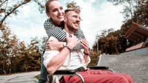 Disabled husband in wheelchair being hugged by his wife standing behind him. Both are smiling, and they are outside.