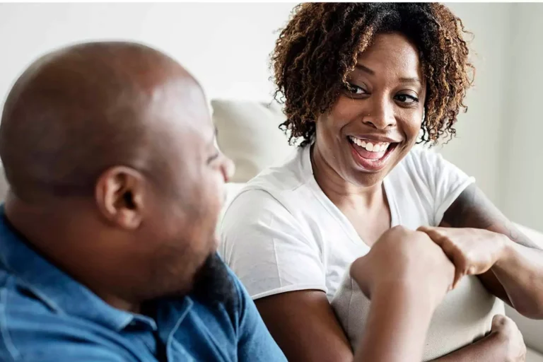 A smiling couple sits together on a couch, engaged in a lively and positive conversation. The woman, wearing a white t-shirt, leans in with a warm and expressive smile, while the man, dressed in a denim shirt, listens attentively. Their body language reflects open communication, illustrating how to give constructive feedback in marriage in a way that strengthens connection and understanding.