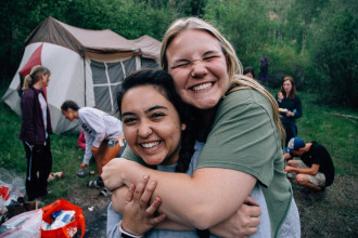 Two smiling teen girls outside at a campsite. One is enthusiastically and tightly hugging the other from behind.