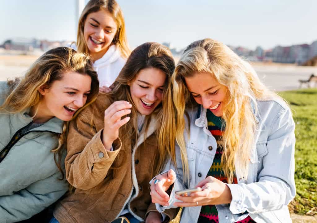 Four teen girls outside laughing together as they look at something on a smart phone