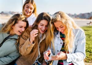 Four teen girls outside laughing together as they look at something on a smart phone