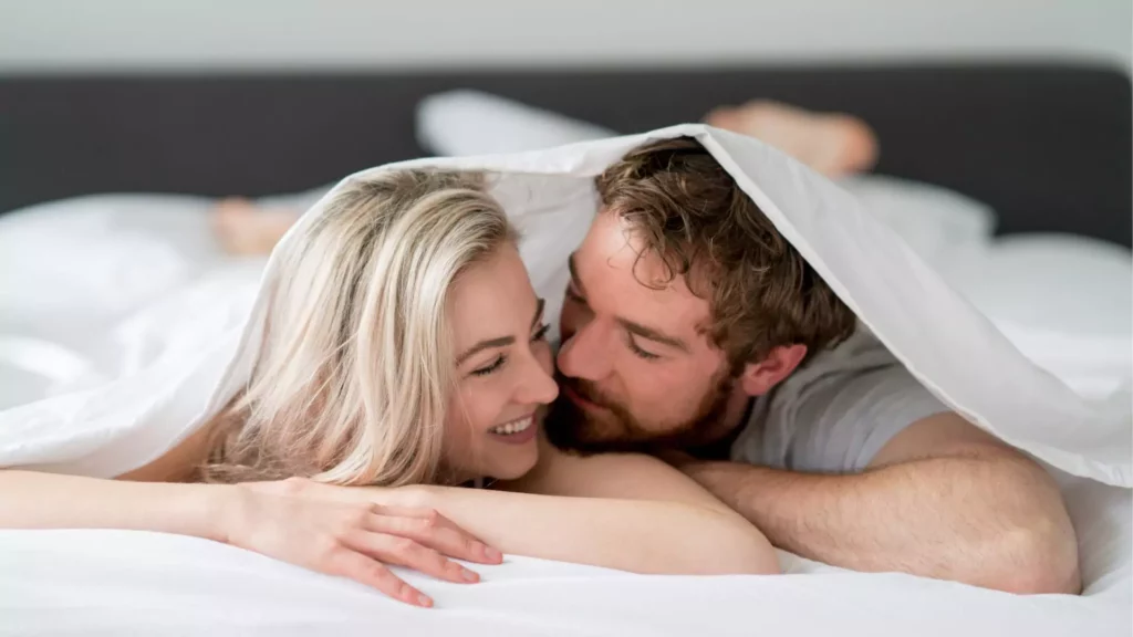 photo of man and woman under blankets in bed. they smile and snuggle intimately because they have a perspective of sex that is healthy.