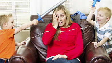 Stressed out mom sitting in a chair as her two young sons hit her with foam toy swords