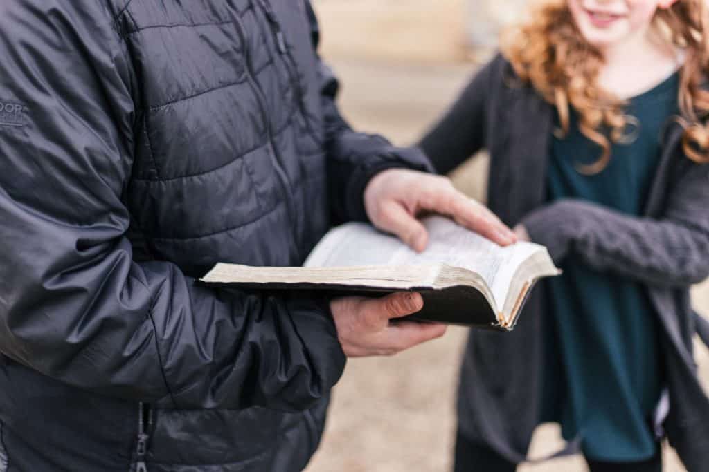 Close up of dad in a winter coat holding an open Bible, with his smiling daughter approaching him in the background